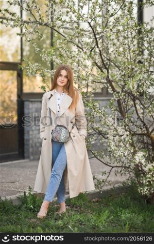 Portrait of a red-haired beauty in the spring nature.. A beautiful girl in a long beige raincoat near a cherry blossom bush 2942.