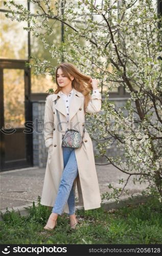 Portrait of a red-haired beauty in the spring nature.. A beautiful girl in a long beige raincoat near a cherry blossom bush 2944.