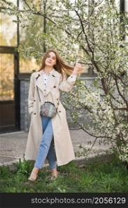 Portrait of a red-haired beauty in the spring nature.. A beautiful girl in a long beige raincoat near a cherry blossom bush 2945.