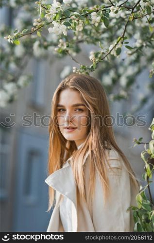 Portrait of a red-haired beauty on the background of a cherry blossom.. A beautiful girl in a long beige raincoat basks in the sun 2972.