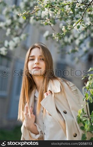 Portrait of a red-haired beauty on the background of a cherry blossom.. A beautiful girl in a long beige raincoat basks in the sun 2973.