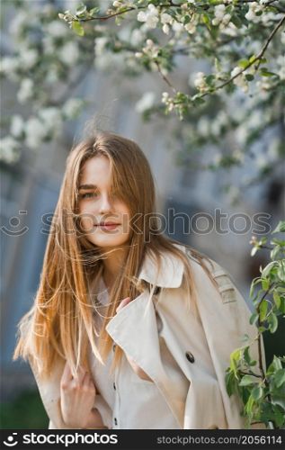 Portrait of a red-haired beauty on the background of a cherry blossom.. A beautiful girl in a long beige raincoat basks in the sun 2974.