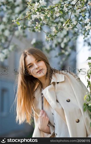 Portrait of a red-haired beauty on the background of a cherry blossom.. A beautiful girl in a long beige raincoat basks in the sun 2976.