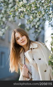 Portrait of a red-haired beauty on the background of a cherry blossom.. A beautiful girl in a long beige raincoat basks in the sun 2976.