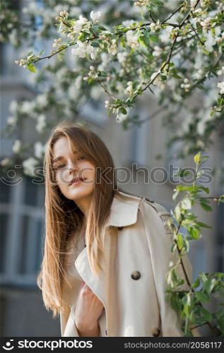 Portrait of a red-haired beauty on the background of a cherry blossom.. A beautiful girl in a long beige raincoat basks in the sun 2977.