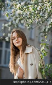 Portrait of a red-haired beauty on the background of a cherry blossom.. A beautiful girl in a long beige raincoat basks in the sun 2977.