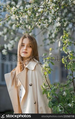 Portrait of a red-haired beauty on the background of a cherry blossom.. A beautiful girl in a long beige raincoat basks in the sun 2978.