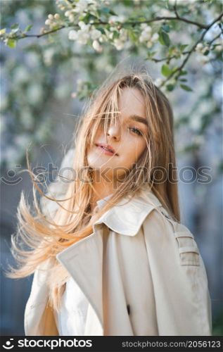 Portrait of a red-haired beauty on the background of a cherry blossom.. A beautiful girl in a long beige raincoat basks in the sun 2982.