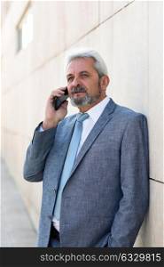 Portrait of a senior businessman talking with a smart phone outside of modern office building. Successful business man wearing blue suit and tie in urban background. Male in formal clothes