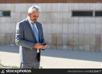 Portrait of a senior businessman texting with a smart phone outside of modern office building. Successful business man in urban background.