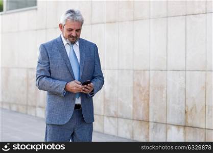 Portrait of a senior businessman texting with a smart phone outside of modern office building. Successful business man in urban background.
