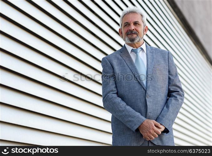 Portrait of a senior businessman with arms crossed outside of modern office building. Successful business man wearing formal suit and tie smiling in urban background.
