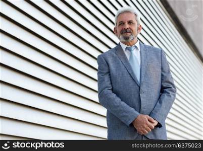Portrait of a senior businessman with arms crossed outside of modern office building. Successful business man wearing formal suit and tie smiling in urban background.