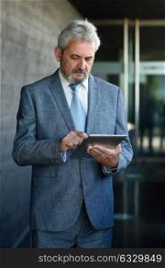 Portrait of a senior businessman with tablet computer outside of modern office building. Successful business man in urban background.
