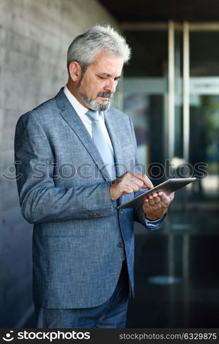 Portrait of a senior businessman with tablet computer outside of modern office building. Successful business man in urban background wearing suit and tie.