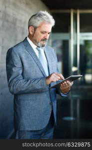 Portrait of a senior businessman with tablet computer outside of modern office building. Successful business man in urban background wearing suit and tie.