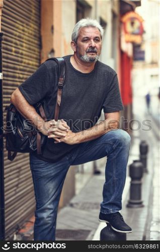 Portrait of a senior tourist man in urban background. Mature male with white hair and beard wearing casual clothes and travel backpack