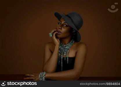 Portrait of a sensual black lady with gorgeous makeup is posing by herself inside a studio wearing a black hat   colorful jewelry.