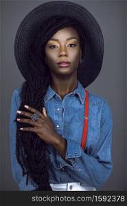 Portrait of a sensual young black female with long dreadlocks & beautiful makeup posing by herself in a studio with grey background wearing colorful jewelry, summer hat, denim shirt & red suspenders.
