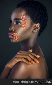 Portrait of a serene young black female with a beautiful short Afro, beautiful makeup, purple lips & orange nails posing by herself in a studio with dark background wearing orange flowers on her face.