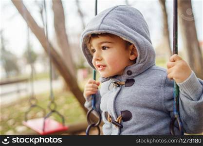 Portrait of a small little cute caucasian boy three years old having fun on the swing in the park in winter or autumn day wearing coat