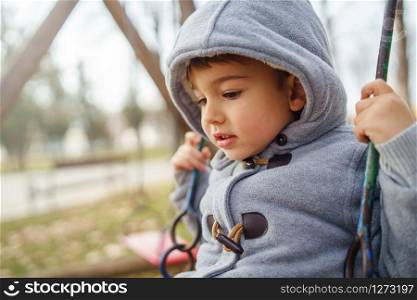 Portrait of a small little cute caucasian boy three years old thinking looking to the ground on the swing in the park in winter or autumn day wearing coat and hood
