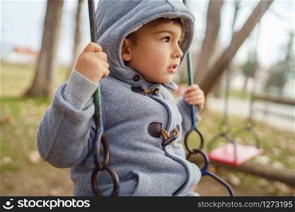 Portrait of a small little cute caucasian boy three years old with hood on the swing in the park in winter or autumn day wearing coat
