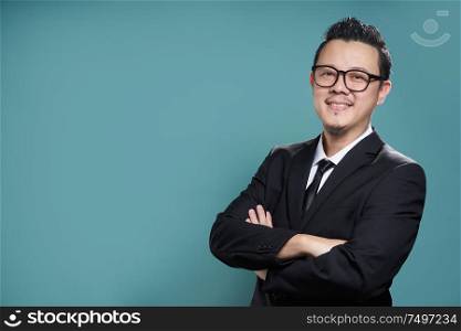 Portrait of a smart asian smiling and confident young businessman in black suit and tie keeping arms crossed , looking at camera isolated on vintage blue color background .