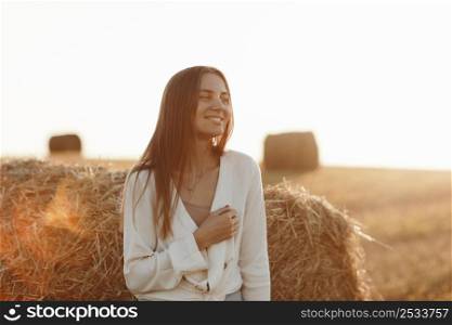 portrait of a smiling beautiful girl with long hair in a jeans skirt. Woman enjoying a walk in a wheat field with hay bales on summer sunny day. portrait of a smiling beautiful girl with long hair in a jeans skirt. Woman enjoying a walk in a wheat field with hay bales on summer sunny day.