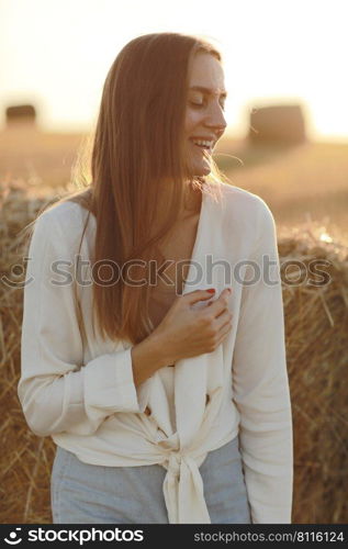 portrait of a smiling beautiful girl with long hair in a jeans skirt. Woman enjoying a walk in a wheat field with hay bales on summer sunny day. portrait of a smiling beautiful girl with long hair in a jeans skirt. Woman enjoying a walk in a wheat field with hay bales on summer sunny day.