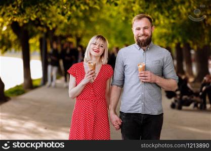 Portrait of a smiling hipsters couple eating ice cream and having fun in the city. stylish young man with beard and blonde woman in red dress are walking in the street.. Portrait of a smiling hipsters couple eating ice cream and having fun in the city. stylish young man with beard and blonde woman in red dress are walking in the street