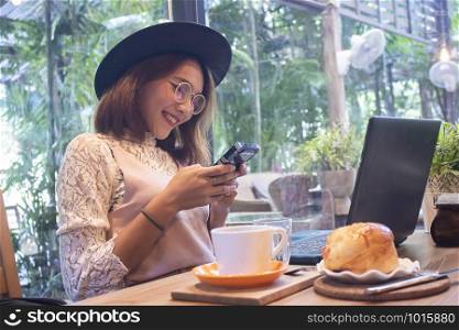 Portrait of a smiling young asian girl holding mobile phone while sitting with laptop computer at a cafe