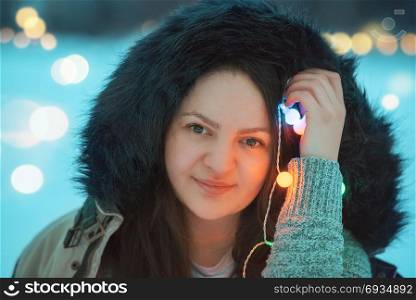 Portrait of a smiling young brunette woman dressed in a winter jacket with a fur hood, holding multicolor string lights near her face.