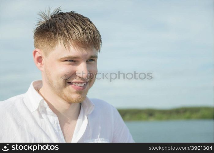Portrait of a smiling young man with a beard wearing a white shirt against the backdrop of a river at summer day