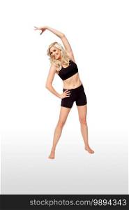 Portrait of a stretching fit young white female athlete with curly long blond hair posing by herself in a studio with a white background wearing black shorts   sports bra.