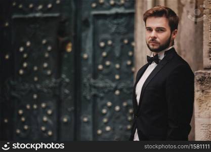 portrait of a stylish young groom in an old European town. man in a stylish suit with bow tie near old building. wedding day.. portrait of a stylish young groom in an old European town. man in a stylish suit with bow tie near old building. wedding day
