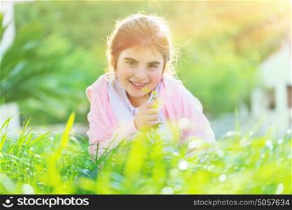 Portrait of a sweet baby girl sitting on the fresh green grass field and enjoying aroma of flower, happy spring holidays in countryside