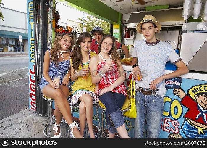 Portrait of a teenage boy standing at a juice counter with two teenage girls and a young woman sitting beside him