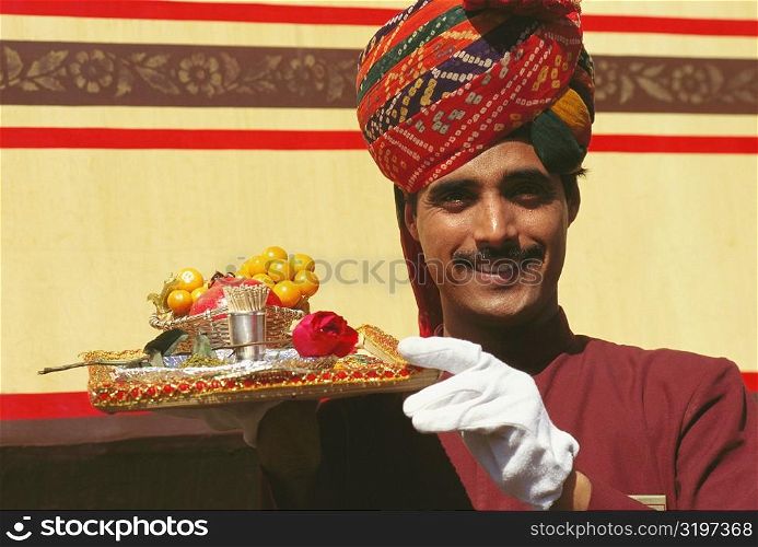 Portrait of a train steward holding winter cherries and a Rose flower on a serving tray, India
