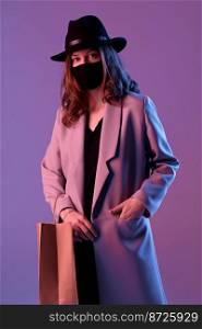 Portrait of a trendy young woman in a coat, hat and protective mask with a paper shopping bag in purple neon lighting.