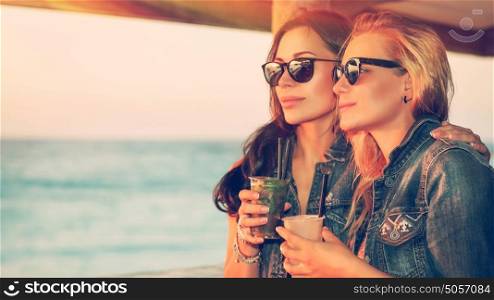 Portrait of a two beautiful girls spending leisure time in the outdoor cafe, drinking tasty cocktails and enjoying beautiful sea view in bright sunny day