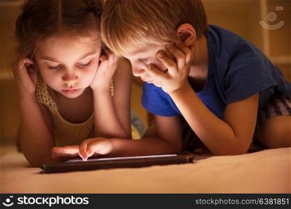 Portrait of a two cute little kids lying down on the floor and watching animated cartoons on the tablet, brother and sister with pleasure spending time together at home
