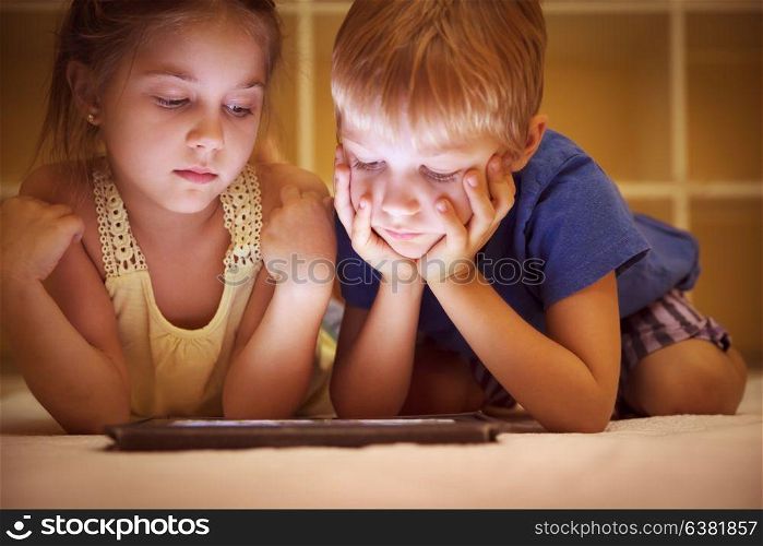 Portrait of a two cute little kids lying down on the floor and watching animated cartoons on the tablet, brother and sister with pleasure spending time together at home