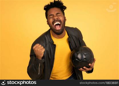 Portrait of a young afro man holding a football ball and celebrating victory while standing against isolated background. Success concept.