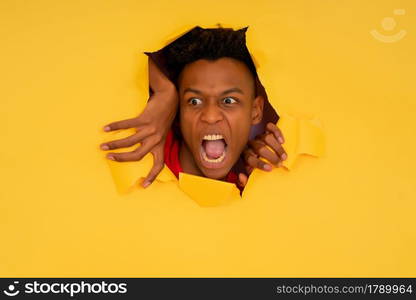 Portrait of a young afro man looking with an angry face through a torn hole in paper wall.