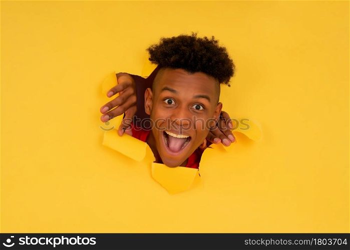 Portrait of a young afro man looking with surprised expression on his face through a torn hole in paper wall.