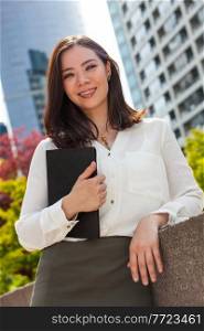 Portrait of a young Asian woman or businessowman smiling in a city holding a folder or tablet computer