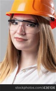 Portrait of a young attractive woman with blond hair in orange helmet and glasses on a neutral gray background.. Portrait of a young attractive woman with blond hair in orange helmet on a neutral gray background.