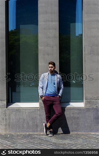 Portrait of a young bearded man, model of fashion, in urban background wearing casual clothes while leaning on a office building wall