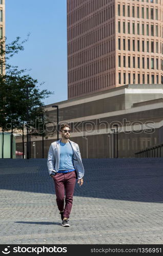 Portrait of a young bearded man with sunglasses, model of fashion, in urban background wearing casual clothes while walking with hand on pocket